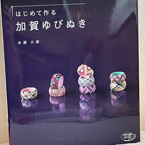 Pode incluir: Um livro intitulado "Como fazer anéis de polegar Kaga" com uma capa roxa e uma foto de anéis de polegar Kaga coloridos sobre uma superfície preta.