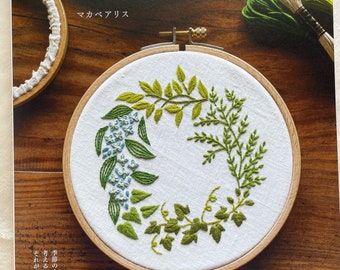 May include: A book cover with a green embroidery hoop featuring a wreath of green leaves and blue flowers. The title of the book is "植物刺繍と 12か月のおはなし." which translates to "Plant Embroidery and 12 Months of Stories." The author is "マカベアリス" which translates to "Macabre Alice."