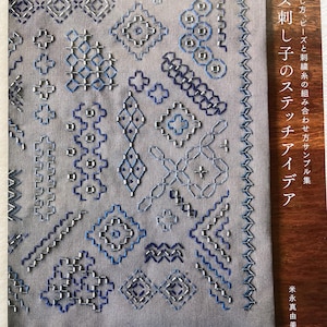Può includere: Una copertina di libro con uno sfondo in tessuto grigio che presenta una varietà di punti di ricamo blu e argentati. Il titolo del libro è "Bead Stitch Ideas" in giapponese. La copertina del libro è in legno.