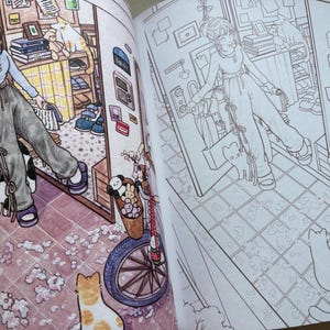May include: An open colouring book displaying two pages with an anime-style illustration of a person in a light blue long-sleeve shirt and dark grey trousers standing in a room with a bicycle, cats, and various household items in both colour and line art.
