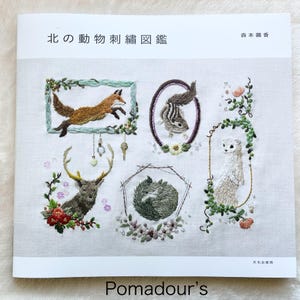 Puede incluir: Un libro titulado "North Animal Embroidery Design" con ilustraciones de animales bordados, incluyendo un zorro, una ardilla, un ciervo y una comadreja. El libro está publicado por "Pomadour's" y presenta un diseño floral.