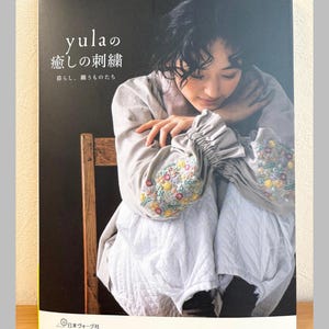 May include: A book cover featuring a woman in a light-colored embroidered outfit. The title "yula's Embroidery" is displayed in Japanese text. The cover also includes Japanese text and a logo for a Japanese publishing company.