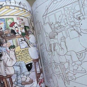 May include: A colouring book open to two pages showing a winter scene with a food truck, a person sitting on a stool holding a mug, a snowman, and a cat. The left page is coloured in soft tones, while the right page features the same line art illustration.