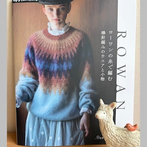 Könnte beinhalten: Buchcover mit dem Titel "ROWAN" und japanischem Text, das eine Frau in einem farbenfrohen Pullover zeigt. Der Pullover hat Farbtöne von Braun, Blau und Rosa. Eine kleine Lama-Figur mit einem Vogel auf dem Rücken befindet sich im Vordergrund.