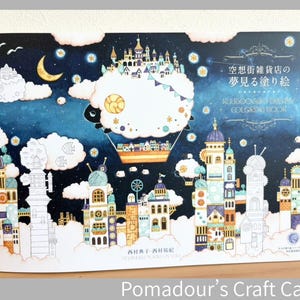 以下が含まれることがあります： 夜空を背景に、白い雲、三日月、星、そして小さな街を運ぶ熱気球が描かれた塗り絵。表紙には「空想街雑貨店の夢見る塗り絵」および「KUUSOOGAI'S DREAMY COLORING BOOK」という文字と、精巧な建物のイラストが印刷されています。