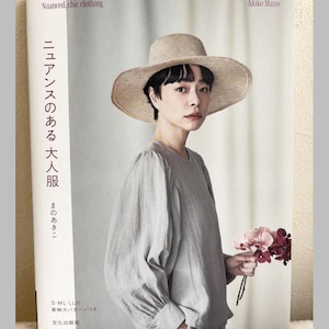Könnte beinhalten: Eine Frau in einer hellgrauen Bluse und einem breitkrempigen Strohhut, die einen Strauß rosa und burgunderfarbener Blumen hält. Das Cover eines Buches mit dem Titel "Nuanced, chic clothing" ist sichtbar, mit Text auf Japanisch. Das Buch ist von Akiko Mano.