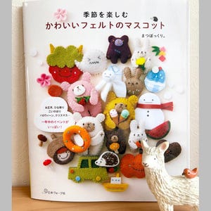 Könnte beinhalten: Ein Buch mit dem Titel "Saisons Spaß, süßes Filz-Maskottchen" mit verschiedenen Filzfiguren. Die Figuren umfassen Tiere, Lebensmittel und saisonale Artikel wie einen Schneemann und einen Kürbis. Eine weiße Lama-Figur mit einem kleinen Vogel befindet sich im Vordergrund.