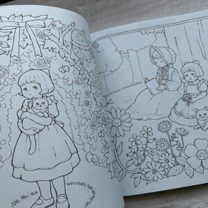 May include: Open book with black and white illustrations. One page shows a girl holding a cat in a garden of roses. The other page depicts two girls and a cat under a tree. Coloring book pages.