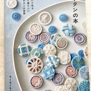 Op de afbeelding: Een boek getiteld "Draadknoopboek" met diverse handgemaakte draadknopen in blauw, wit en roze. De knopen zijn gerangschikt op een lichtgrijze plaat. Het boek heeft Japanse tekst.