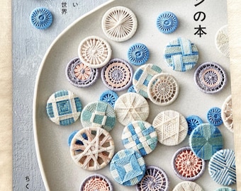 Puede incluir: Un libro titulado "Libro de botones de hilo" con varios botones de hilo hechos a mano en azul, blanco y rosa. Los botones están dispuestos sobre un plato gris claro. El libro tiene texto japonés.