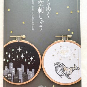 May include: A book titled "Sparkling Starry Sky Embroidery" features two embroidery hoops with night sky and whale designs. The cover is dark grey with gold stars and Japanese text. The book is by X-Knowledge and Pomadour's.