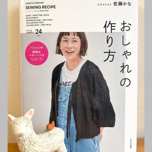 May include: A book titled "KANA'S STANDARD SEWING RECIPE" with a woman on the cover. The book includes patterns for shirts, pants, blouses, and skirts. A white llama figurine with a small orange bird on its back is in the foreground.