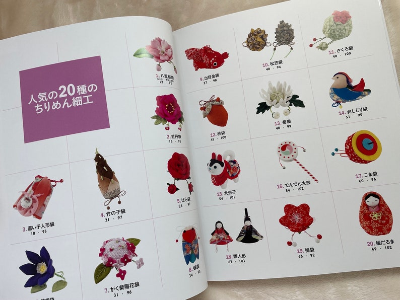 May include: A book with a pink cover featuring 20 different types of Japanese crafts made with fabric. The crafts are displayed in a grid format with a Japanese name and number for each craft. The book title is "Popular 20 Types of Chirimen Crafts".