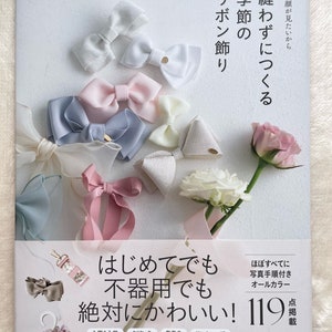 以下が含まれることがあります： 表紙にピンクと白のリボンが付いた本の表紙。本のタイトルは「縫わずにつくるリボン飾り」。副題は「季節の飾り」。本の表紙には「初心者でも作れます！119デザイン」という文字も書かれています。