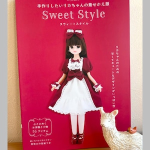 Op de afbeelding: Een rood boek met de titel "Sweet Style" met Japanse tekst en een poppenillustratie. De pop draagt een bordeauxrode en witte jurk. Rechts staat een decoratief lamafiguur.