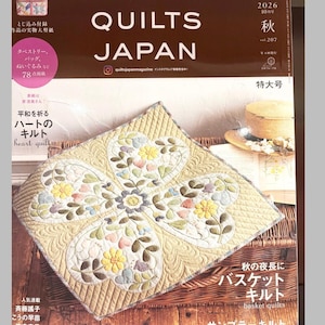 Puede incluir: La edición de octubre de 2026 de la revista Quilts Japan, volumen 207, presenta en la portada una pieza acolchada cuadrada con un patrón floral de cuatro corazones en tonos pastel. La portada incluye texto en japonés, un sombrero y una cesta.