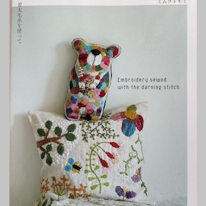 Può includere: Una copertina di libro con un orso ricamato colorato e un cuscino con ricami floreali. Il testo recita "Embroidery sewed with the darning stitch" e "POMADOUR'S". L'orso è sopra il cuscino, mostrando la tecnica del rammendo.