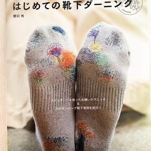 May include: A book titled "はじめての靴下ダーニング" (Hajimete no Kutsushita Darning) by Hikaru Noguchi, featuring a close-up of two socks with colorful embroidery. The book cover also includes the text "野口光" (Noguchi Hikaru) and "Pomadour's Craft Cafe".