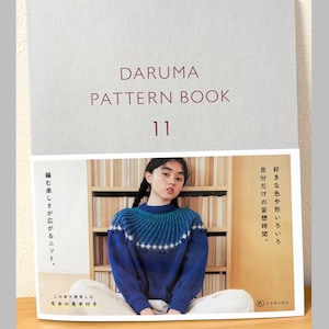 以下が含まれることがあります： DARUMA PATTERN BOOK 11と書かれたライトグレーの表紙のソフトカバー本。表紙には、ターコイズブルーと白のヨーク模様が入った青いニットセーターを着た人物の写真が印刷されており、背景には木製の本棚が写っている。