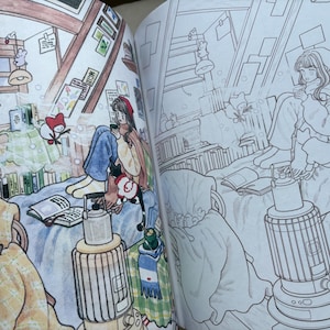 May include: A colouring book spread featuring two side-by-side illustrations of an attic room. One side is coloured with warm tones, showing a person sitting on a bed with a book, a heater, and a cat. The other side is a black and white line drawing.