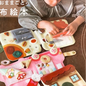 May include: A felt book with different scenes, including a kitchen, a bakery, and a house. The book is open and a child is playing with it. The book is titled "Felt Book" in Japanese.