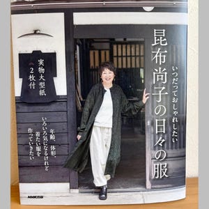 以下が含まれることがあります： 白いシャツと白いパンツの上に、ダークグリーンの柄入りロングコートを羽織った人物が写る書籍の表紙。表紙には日本語のテキスト、黒い衣服のグラフィック、NHK出版のロゴが、木造建築の背景とともにデザインされています。