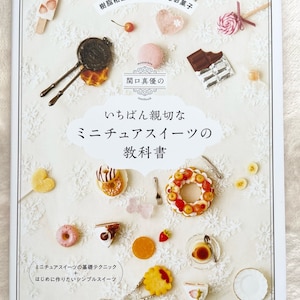 以下が含まれることがあります： 白い背景に、ドーナツ、ケーキ、キャンディーなど、さまざまなミニチュアのお菓子が載っている本の表紙。本のタイトルは「いちばん親切な ミニチュアスイーツの 教科書」で、著者は「関口真優」です。