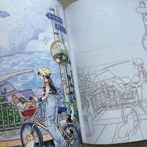 May include: An open colouring book featuring two side-by-side illustrations of a person with a bicycle near a coastal town. The left page is coloured in watercolour tones, while the right page shows the same scene in black and white line art.