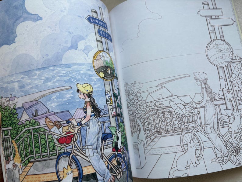 May include: An open colouring book featuring two side-by-side illustrations of a person with a bicycle near a coastal town. The left page is coloured in watercolour tones, while the right page shows the same scene in black and white line art.