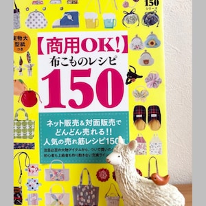 Może przedstawiać: Żółta książka zatytułowana "Przepis na wyroby z tkanin 150" z japońskim tekstem i ilustracjami toreb i akcesoriów. Biała figurka lamy z małym pomarańczowym ptakiem na pierwszym planie.