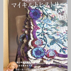 Könnte beinhalten: Ein japanischsprachiges Buch mit dem Titel "my quilt history", das ein detailliertes Bild eines Quilts mit Blumen- und Blättermustern in Lila, Blau und Grün zeigt. Das Buch befindet sich neben einer kleinen Hundefigur auf einem Regal.