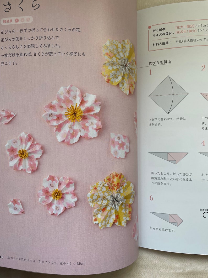May include: A step-by-step guide on how to fold origami cherry blossoms. The instructions are written in Japanese. The image shows a close-up of the folded paper flowers, with the finished product on the left and the folding steps on the right. The flowers are made from pink and yellow paper with white polka dots.
