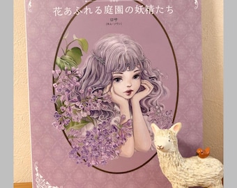 Puede incluir: Un libro para colorear japonés, "Hadas del jardín lleno de flores", con una portada lila y una ilustración de una mujer y flores. Junto a él, una figura de llama blanca con un pájaro naranja. El libro está sobre una superficie de madera.