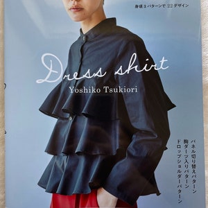Könnte beinhalten: Ein Buch mit dem Titel "Dress Shirt" von Yoshiko Tsukiori, das ein schwarzes Hemd mit Rüschenkragen und ein Model zeigt, das das Hemd trägt. Der Buchumschlag enthält auch den Text "月居良子のドレスシャツ 身頃3パターンで22デザイン" und "7-9-11-13-15号 実物大型紙つき 主婦と生活社".