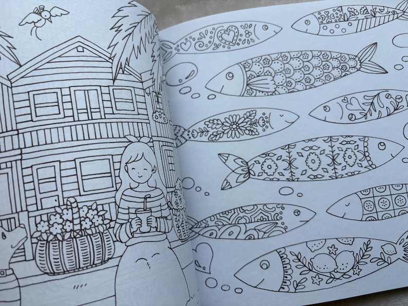 May include: An open coloring book page with detailed black and white illustrations. One page features a house, a girl holding a drink, and a dog. The other page displays several fish with intricate patterns, including hearts, flowers, and geometric designs.