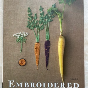以下が含まれることがあります： 「Embroidered Kitchen Garden」というタイトルと「Pomadour's Craft Cafe」というサブタイトルが書かれた本の表紙。表紙には、刺繍されたニンジン、パセリ、花が描かれている。
