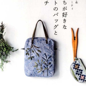 Op de afbeelding: Een blauwe quilted tote bag met een bloemenpatroon en bruine leren handvatten. De tas hangt aan een witte muur met een kleine blauwe en witte quilted pouch ernaast.