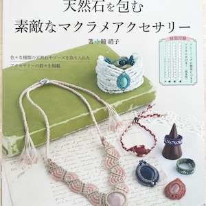 Może przedstawiać: Okładka książki z różowo-białym naszyjnikiem z makramą z kamieniem różowym kwarcem, niebieską bransoletką z makramą z turkusem i czerwoną bransoletką z makramą z czerwonym kamieniem. Okładka książki ma tytuł "Piękne akcesoria z makramą owinięte naturalnymi kamieniami" w języku japońskim.