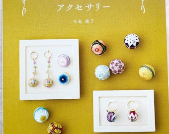 May include: A book titled "野の花の小さなてまりとアクセサリー" by 寺島綾子. The book cover features a yellow background with a floral design and several small, colorful, embroidered balls displayed on white trays. The text "Pomadour's Craft Cafe" is printed at the bottom of the cover.