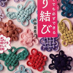 Op de afbeelding: Een verzameling kleurrijke, ingewikkeld geknoopte decoratieve elementen in verschillende vormen en maten. De knopen zijn in tinten blauw, groen, rood, paars en goud, gerangschikt op een roze achtergrond. De afbeelding bevat ook Japanse tekst.