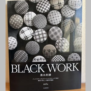 May include: A book titled "BLACK WORK" with various black and white embroidered circular designs. The book cover features the title in large white letters, with Japanese text below. The book is on a wooden surface.