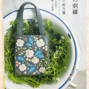Puede incluir: Un libro titulado "yula no 刺繍" con una descripción en japonés en la portada. El libro presenta una imagen de primer plano de una pequeña bolsa de mano gris con un diseño de bordado floral. La bolsa está sentada en un cuenco de cerámica blanco lleno de plantas verdes.