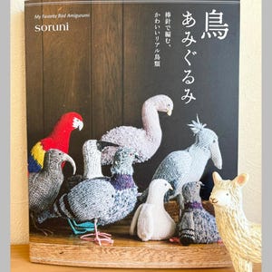 May include: A book titled "My Favorite Bird Amigurumi" by soruni, featuring various knitted bird amigurumi. The cover displays a collection of colorful, handcrafted birds. A small llama figurine is in the foreground. The text "Pomadour's Craft" is at the bottom.