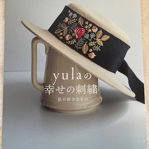 May include: A book titled "yula's Happy Sewing: My Favorite Things" with a beige hat with a black ribbon and embroidered flowers and mushrooms on the ribbon. The hat is resting on a beige ceramic mug.