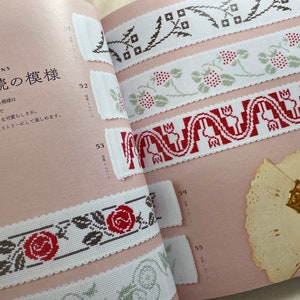 May include: A book with a pink cover and white pages showing different embroidery patterns. The patterns are for borders and feature flowers, leaves, and geometric shapes. The book is titled "こ方連続の模様" which translates to "Continuous Patterns".