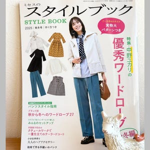 Peut inclure: Une couverture de magazine avec du texte japonais, intitulée "STYLE BOOK 2025". Elle présente une femme en veste blanche et pantalon bleu, avec des images de vêtements. Une figurine décorative de lama est au premier plan. Le texte "Pomadour's Craft" est visible.