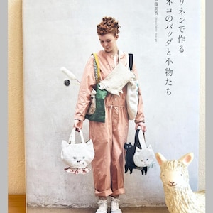Könnte beinhalten: Eine Frau hält katzenförmige Tragetaschen und ein Buch mit dem Titel "Katzen-Taschen und Accessoires aus Leinen herstellen". Die Taschen sind weiß und schwarz, mit Katzen-Gesichtern. Die Frau trägt einen rosa Overall und weiße Turnschuhe. Im Hintergrund ist ein dekoratives Lama.