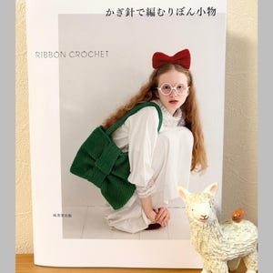 以下が含まれることがあります： 「リボンかぎ針編み」というタイトルの本。赤いリボンとメガネをかけた女性が、緑のかぎ針編みバッグを持っています。オレンジ色の鳥が付いた白いラマの置物も近くにあります。本は木製の表面にあります。