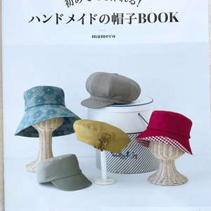 May include: A book titled "Handmade Hat Book" with the subtitle "Even beginners can make it!" by mameco. The book cover features five different hats in various colours and styles, including a red and white gingham hat, a green floral hat, a brown newsboy cap, a yellow beret, and a green baseball cap.