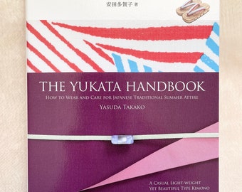 May include: The cover of a book titled "The Yukata Handbook: How to Wear and Care for Japanese Traditional Summer Attire" by Yasuda Takako. The book cover features a red and white striped pattern with a blue stripe. The title is in white text on a purple background. The book is published by Seibun-do Shinkosha.